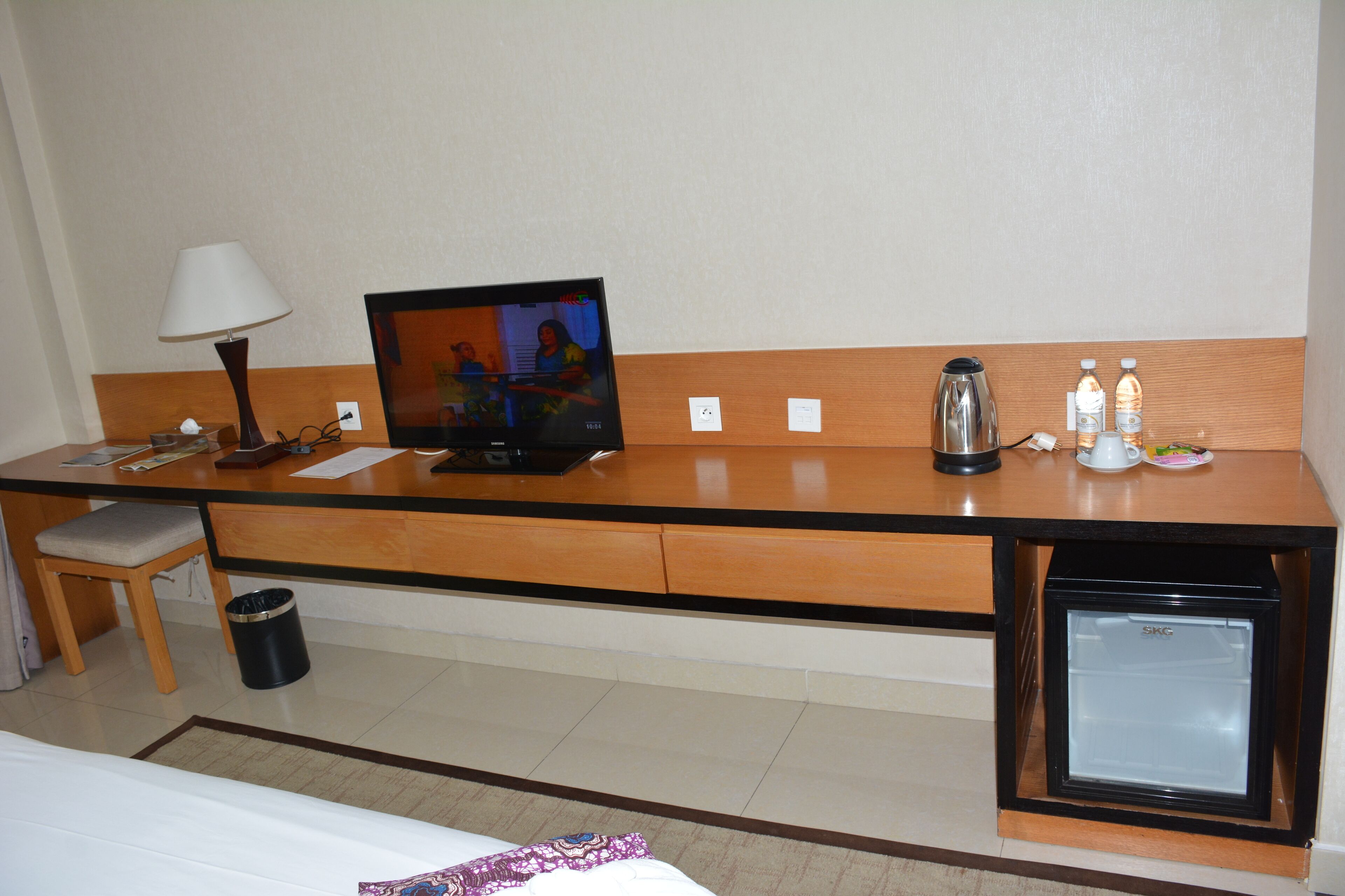 Ein Hotelzimmer mit einem Holztisch, einem Fernseher, einer Kaffeemaschine, einem Minikühlschrank, einem Mülleimer und einem Bett.