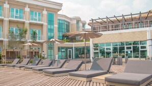 Un área de lounge en la azotea con varios sillones de sol y sombrillas, una terraza de madera y un edificio moderno con ventanas grandes y balcones.