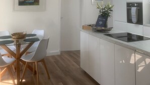 Una cocina moderna con armarios blancos, horno empotrado y comedor con mesa de vidrio y sillas blancas.