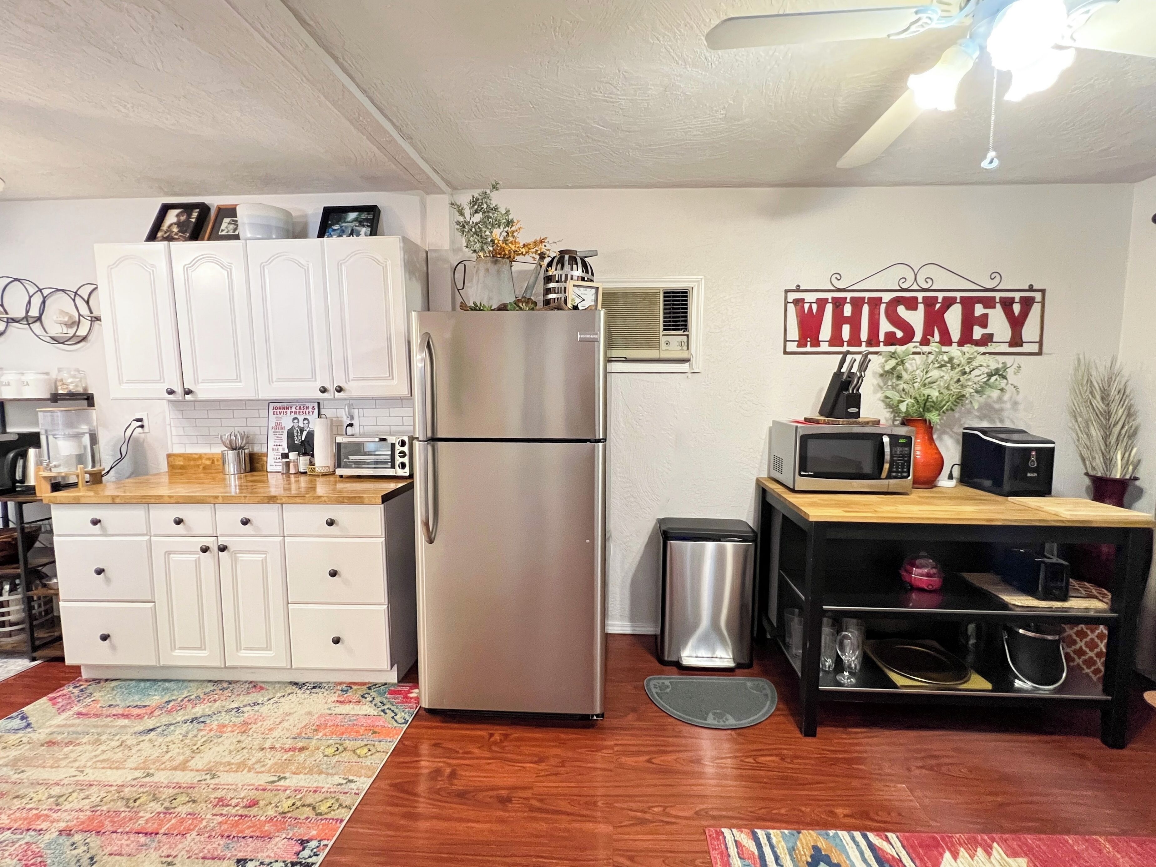 A compact kitchen with white cabinets, a stainless steel refrigerator, a microwave, and a toaster. A 'WHISKEY' sign is visible on the wall.