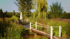 Eine Holzbrücke über einen kleinen Teich, umgeben von üppiger Vegetation und einem Picknickplatz mit Stühlen und Sonnenschirm.