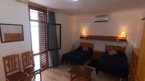 Una habitación con dos camas, una mesa, una silla, un ventanal con persianas, un ventilador de techo y un equipo de aire acondicionado.