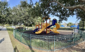 Explore the playground area, featuring a play structure with slides and climbing elements, surrounded by trees and a green fence.
