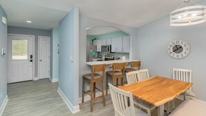 A modern kitchen with white cabinets, a central island, and a dining area with a wooden table and white chairs. - Perfect Private Navarre Beach Getaway! 4 minute from the beach! (Navarre)