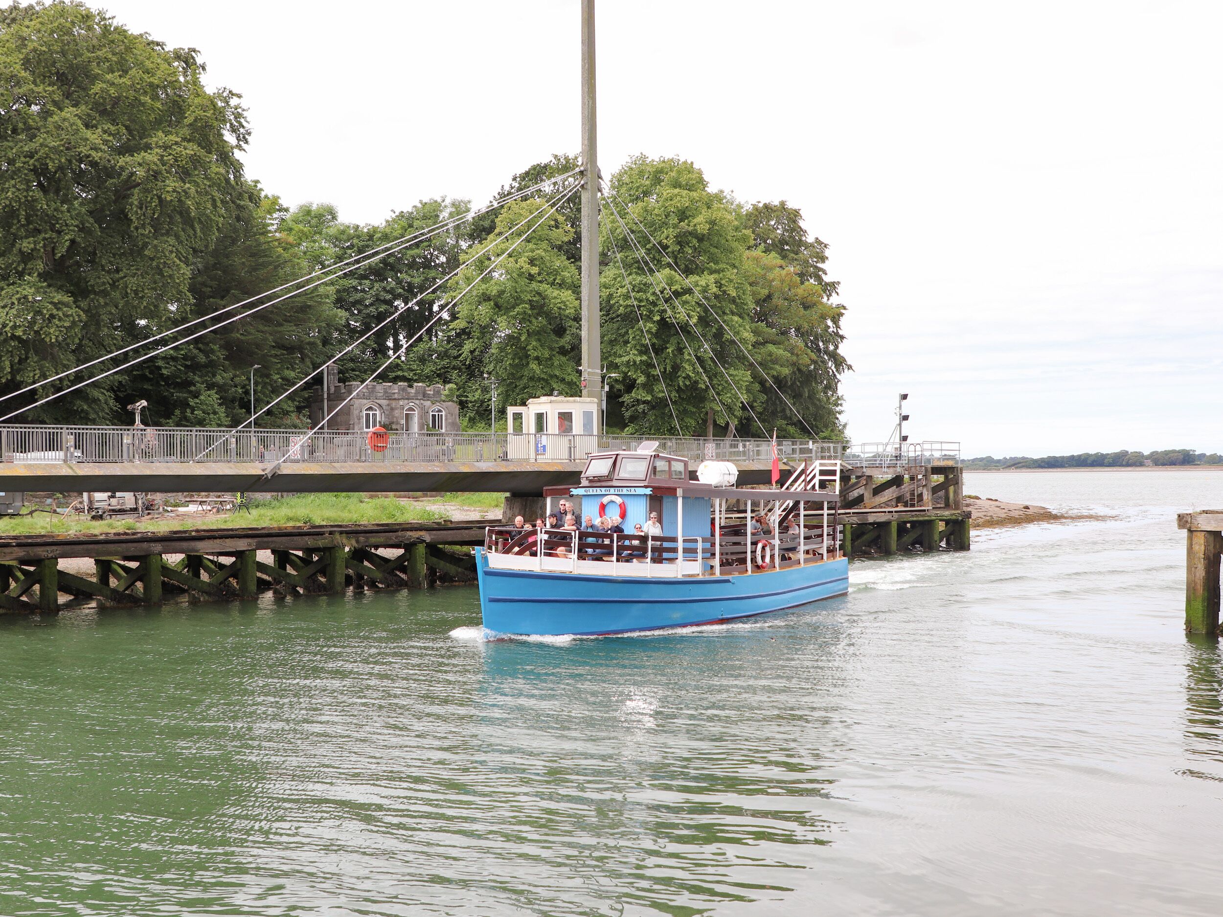 A blue boat with passengers, docked at a pier with a bridge overhead.