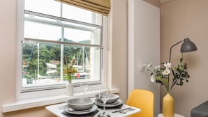 A dining area with a table set for two, a yellow chair, a small side table with a lamp, and a view of a marina through a window. - Cwm Gwyrfai (Caernarfon)