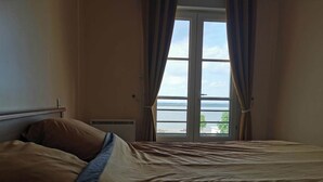 A bedroom with a large bed, a window with curtains, and a view of the sea. - Maison Dans Charmante Bourgade (Paimbœuf)