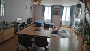 A modern living room with a wooden dining table, black chairs, a sofa, and a coffee table. There is a bookshelf, a desk, and a hanging plant. - Maison Dans Charmante Bourgade (Paimbœuf)