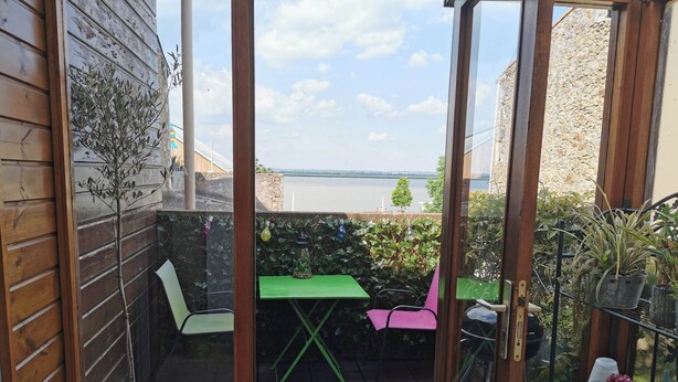A small patio with a wooden table and chairs, a potted plant, and a view of a body of water. - Maison Dans Charmante Bourgade (Paimbœuf)