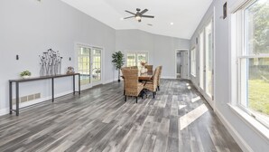 A spacious room with a dining area featuring a wooden table and wicker chairs, a ceiling fan, and large windows allowing natural light. - 4BR, 4BA, Amazing Indoor Pool, EV charger, ~10 acres retreat, next to Park (Big Pool)