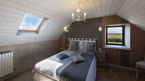 A bedroom with a sloped ceiling, a bed with a headboard, two bedside tables, a nightstand, a radiator, and two windows with views of the outdoors.