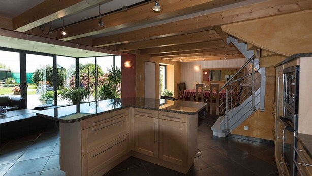 A spacious kitchen with a large island, wooden beams on the ceiling, and a view of a garden through large windows.