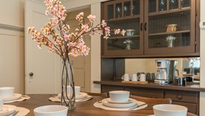 A dining table set with white plates and bowls, a vase with pink flowers, and a kitchen in the background with cabinets and appliances.