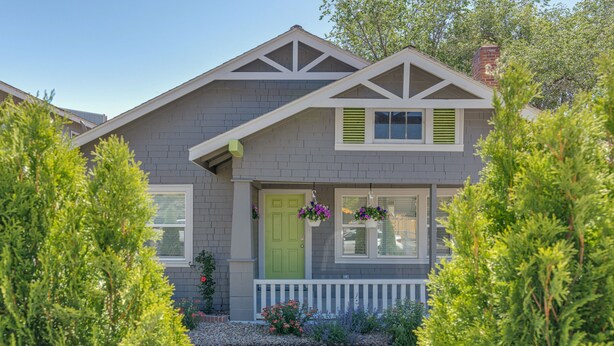 A two-story house with a front porch, a green door, and a small garden with flowers and shrubs.