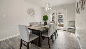 En matplats med ett träbord och stolar, en kristallkrona, en krukväxt och utsikt över utomhusmiljön genom en glasdörr.