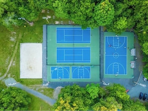 An aerial view of a sports complex with multiple tennis courts and a basketball court, surrounded by dense greenery.