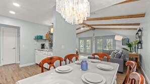 Comedor con mesa blanca para cuatro, sillas de madera, candelabro y cocina al fondo.