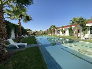 Una piscina con sillas de descanso, rodeada de palmeras y un edificio con ventanas arqueadas.