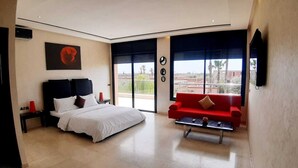 Een moderne hotelkamer met een groot bed, een rode bank, een bijzettafeltje en uitzicht op de stad door grote ramen.
