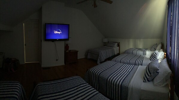 A hotel room with a television displaying the Disney+ logo, two single beds with striped bedding, a wooden headboard, and a small bedside table with a lamp.