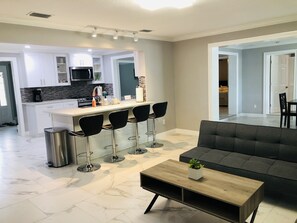 A modern living room with a grey sofa, a wooden coffee table, and a kitchen area with white cabinets, a black countertop, and a black chair. - Cozy Home 4 Minutes From Indian Rocks Beach (Largo)