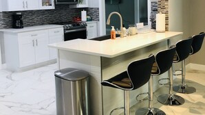 Cuina moderna amb armaris blancs, un vàter d'acer inoxidable i un terra de marbre. Hi ha tres tamborets negres i un cubell d'escombraries d'acer inoxidable.