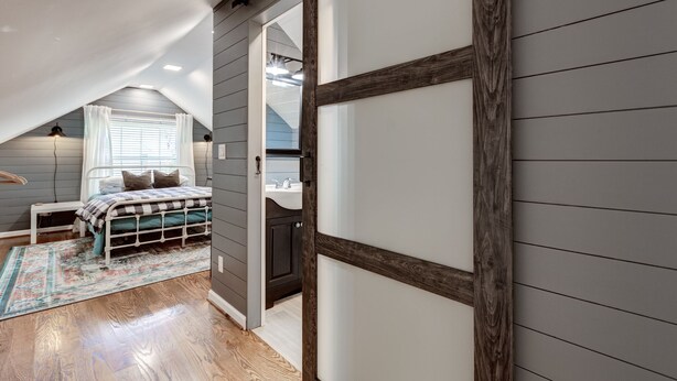 A bedroom with a sloped ceiling, a bed with a checkered bedspread, a bedside table, a lamp, and a window with curtains. - Deacon Stays: The Idlewilde - Walk to WFU (Winston-Salem)