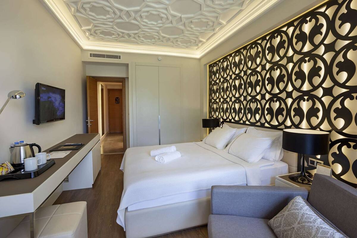 A hotel room with a large bed, a desk with a flat-screen TV, a coffee maker, and a decorative wall panel.