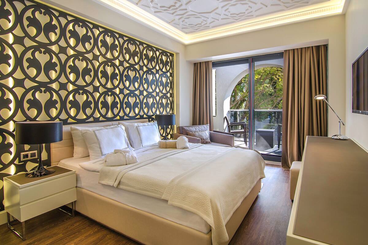 A modern hotel room with a large bed, a nightstand, a desk, a TV, and a balcony with a view of trees.