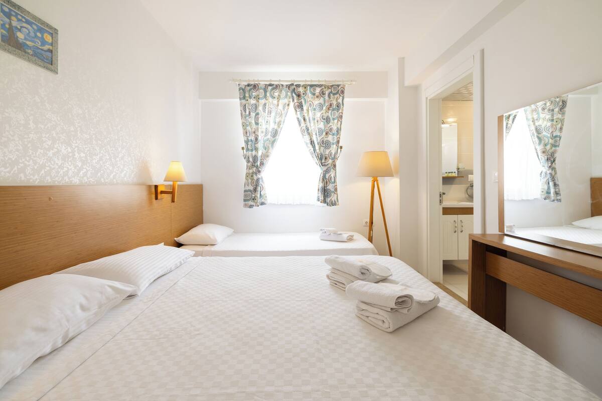 Habitación de hotel con dos camas, cabecero de madera, mesita de noche con lámpara, espejo y ventana con cortinas estampadas.
