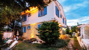 A two-story house with a stone facade, blue shutters, and a well-maintained garden with a pathway leading to the entrance. - Casa Gaia Otel (Bozcaada)