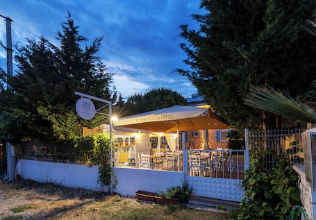 A cozy outdoor seating area with tables and chairs under a canopy, surrounded by trees and a white wall.