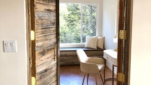 A room with a wooden door, a white chair, a desk, and a window with a view of trees. - Architectural Peace and Perfection in the Sandia Mountains (Albuquerque)