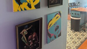 Une pièce avec quatre œuvres d’art encadrées accrochées au mur, un coin repas avec des chaises et une table, et une poubelle.