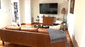En stue med en lædersofa i brunt, et fladskærms-tv, en reol og en loftsvifte.