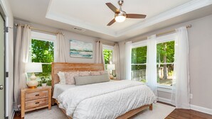 Une chambre à coucher avec un grand lit, une table de chevet, un ventilateur de plafond et une fenêtre avec des rideaux.