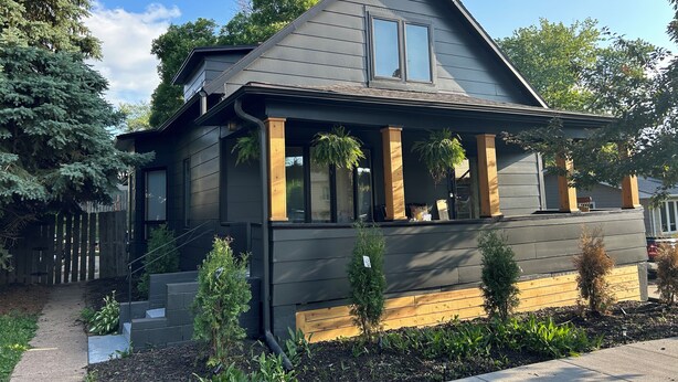 Ein modernes, zweistöckiges Haus mit dunkler Fassade, einer überdachten Veranda mit Holzsäulen und einem kleinen Gartenbereich mit verschiedenen Pflanzen.