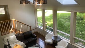Ein geräumiges Wohnzimmer mit hoher Decke, Oberlichtern, einer Treppe, einem Ledersofa, einem weißen Sessel, einem kleinen runden Tisch und einer Lampe.