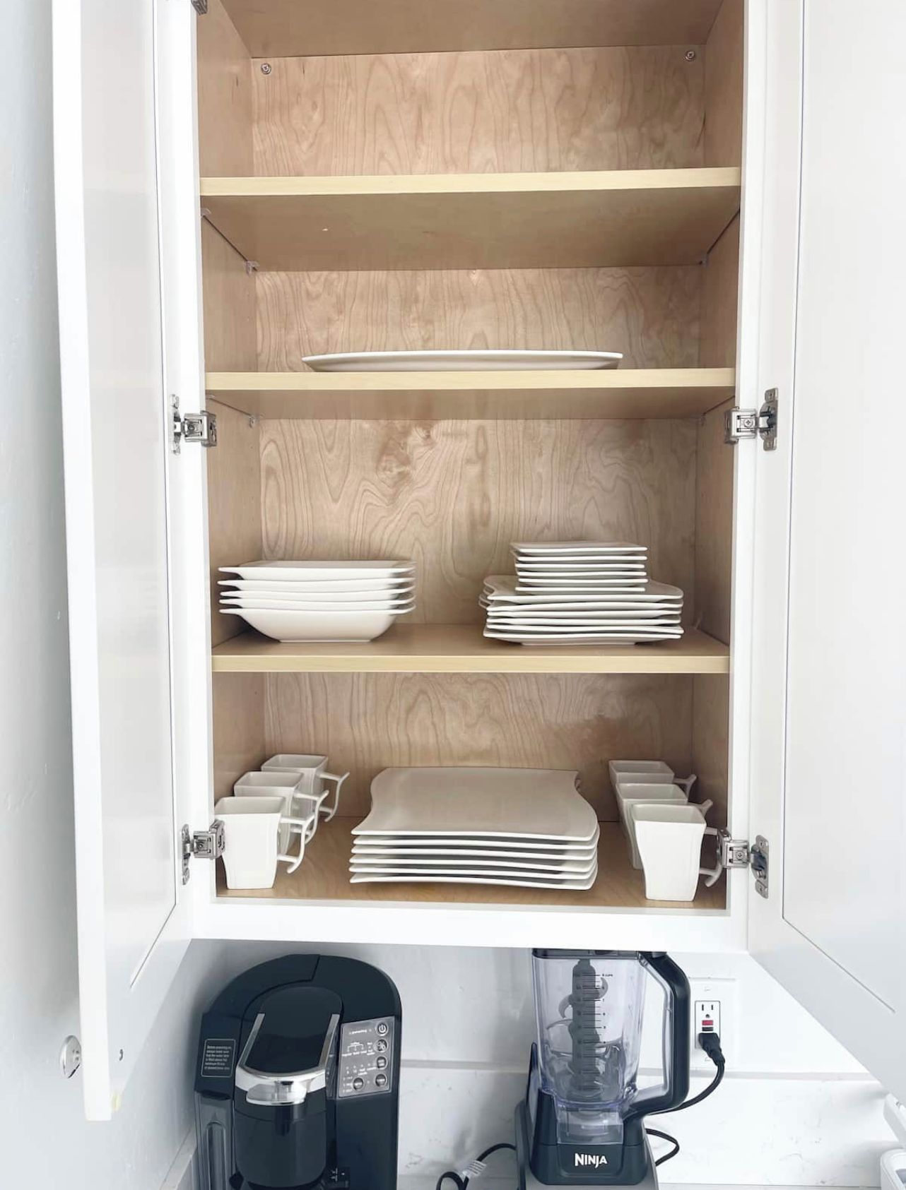 A kitchen cabinet with shelves holding white plates, bowls, and cups. Below, there are two coffee makers and a blender.