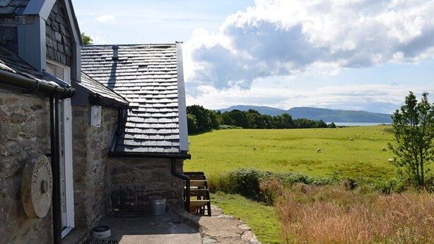 A stone building with a sloped roof, a small outdoor seating area, and a view of a green field with sheep. - Restored watermill with stunning views of Loch Fyne (Kilfinan)