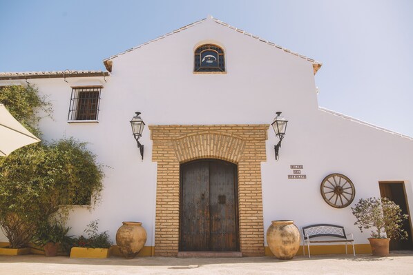 A white building with a large wooden door, two wall-mounted lanterns, and a sign above the door. - Villa con Vistas Espectaculares en Malaga (Casabermeja)