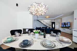 A modern dining area with a round table set for six, a decorative light fixture above, and a staircase in the background.