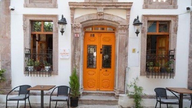 A building with a prominent orange door, two windows with metal railings, and a cobblestone street. - Alancha Otel (Ayvalik)