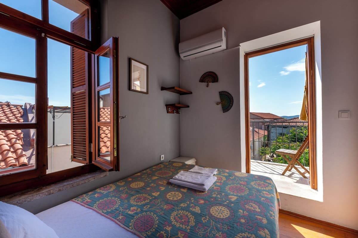 Un dormitorio con cama, ventana con persianas de madera, unidad de aire acondicionado y balcón con una silla.