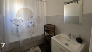Ein Badezimmer mit Duschvorhang mit floralem Muster, einem weißen Waschbecken mit Spiegel darüber und einem Holzregal.