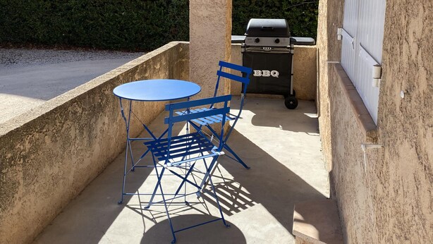 Eine überdachte Terrasse im Freien mit einem blauen Tisch und Stühlen, einem Grill und Blick auf ein Wohngebäude im Hintergrund.