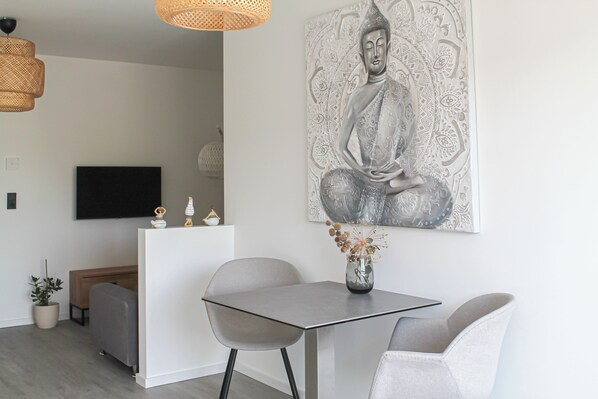A modern living room with a minimalist design, featuring a dining table, two chairs, a sofa, a TV, and a large Buddha artwork on the wall.