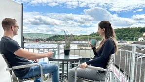 Deux personnes sont assises autour d’une table ronde, près d’un pot de fleurs, et elles ont devant elles une vue sur un paysage urbain avec des bâtiments et de la verdure.