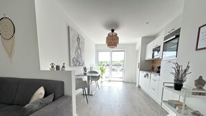 Un espace de vie moderne avec un canapé gris, un coin repas comprenant une table et des chaises, une cuisine avec des meubles blancs et une vue sur l’extérieur, visible à travers une porte vitrée.