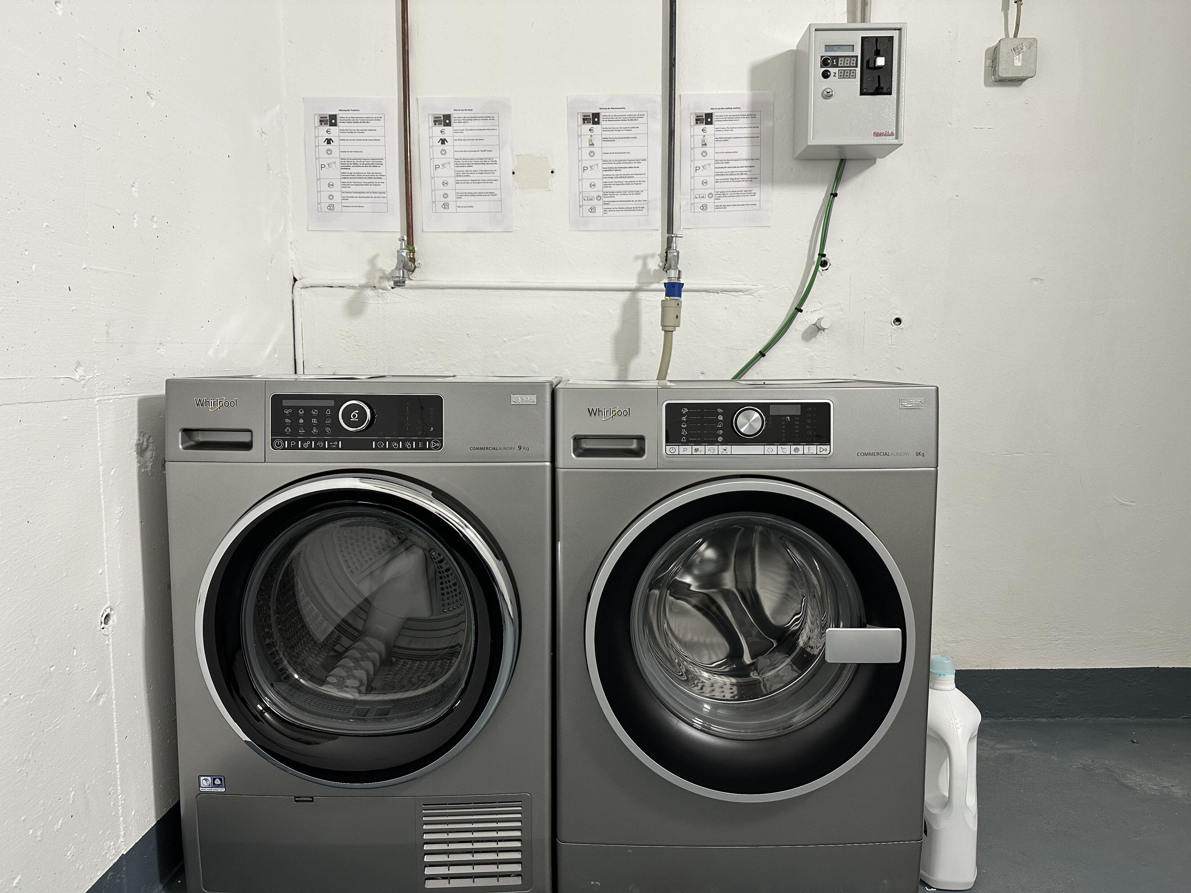 Kaksi Whirlpool-merkkiä olevaa, edestä täytettävää pyykinpesukonetta, joissa on ohjauspaneelit, seinälle asennettu sähkörasia ja lattialla oleva vesipullo.